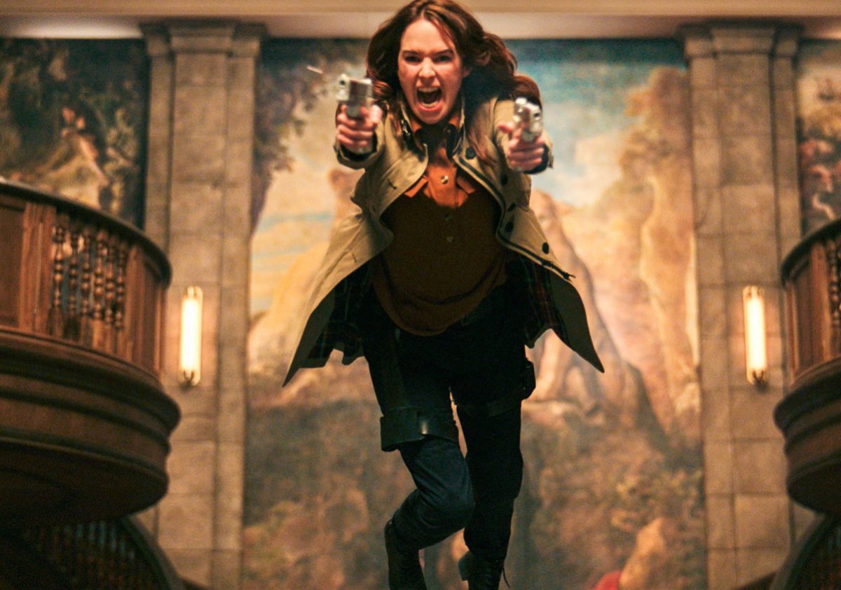 Lena Headey comes over a desk screaming with guns in both hands in Gunpowder Milkshake