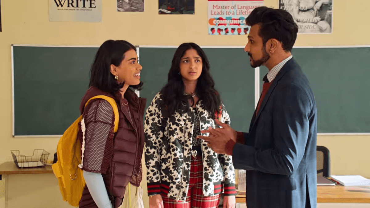 Aneesa smiles as she talks to Mr. Kulkarni in his classroom, one of the school's teachers who is also South Asian. Devi stands behind them, frowning with a look of betrayal on her face as she looks at Mr. Kulkarni.