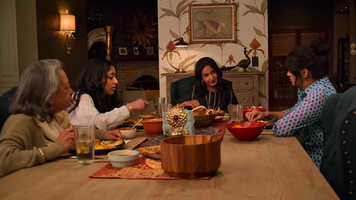 The Vishwakumar family sits around a table eating dinner. Nalini sits at the head of the table and is speaking with Kamala, to her left. Devi sits on Nalini's right and Nirmala is in the foreground of the image, next to Devi. The family is eating a traditional Indian meal prepared by Nirmala.