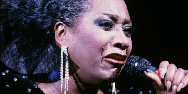 Patti LaBelle performs in the 1980s, crying with emotion into the microphone. she has on long dangly, sparkly earrings, bold make up, long nails, and her signature big hair pulled back.