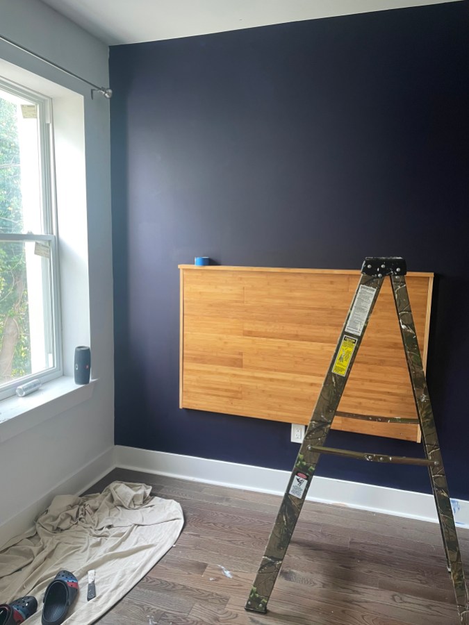 A dark purple wall with a wooden headboard mounted on the wall, a ladder and a pair of Crocs in the foreground