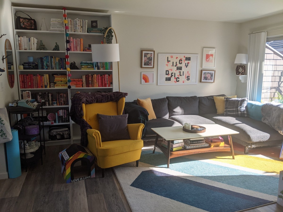 Large bookcases fill the left side of the frame, filled with books organized by color, and colorful prints are on the wall over a dark couch. A yellow armchair sits next to the couch along with a a grey and gold coffee table, and a white and blue area rug sits on the floor over grey flooring