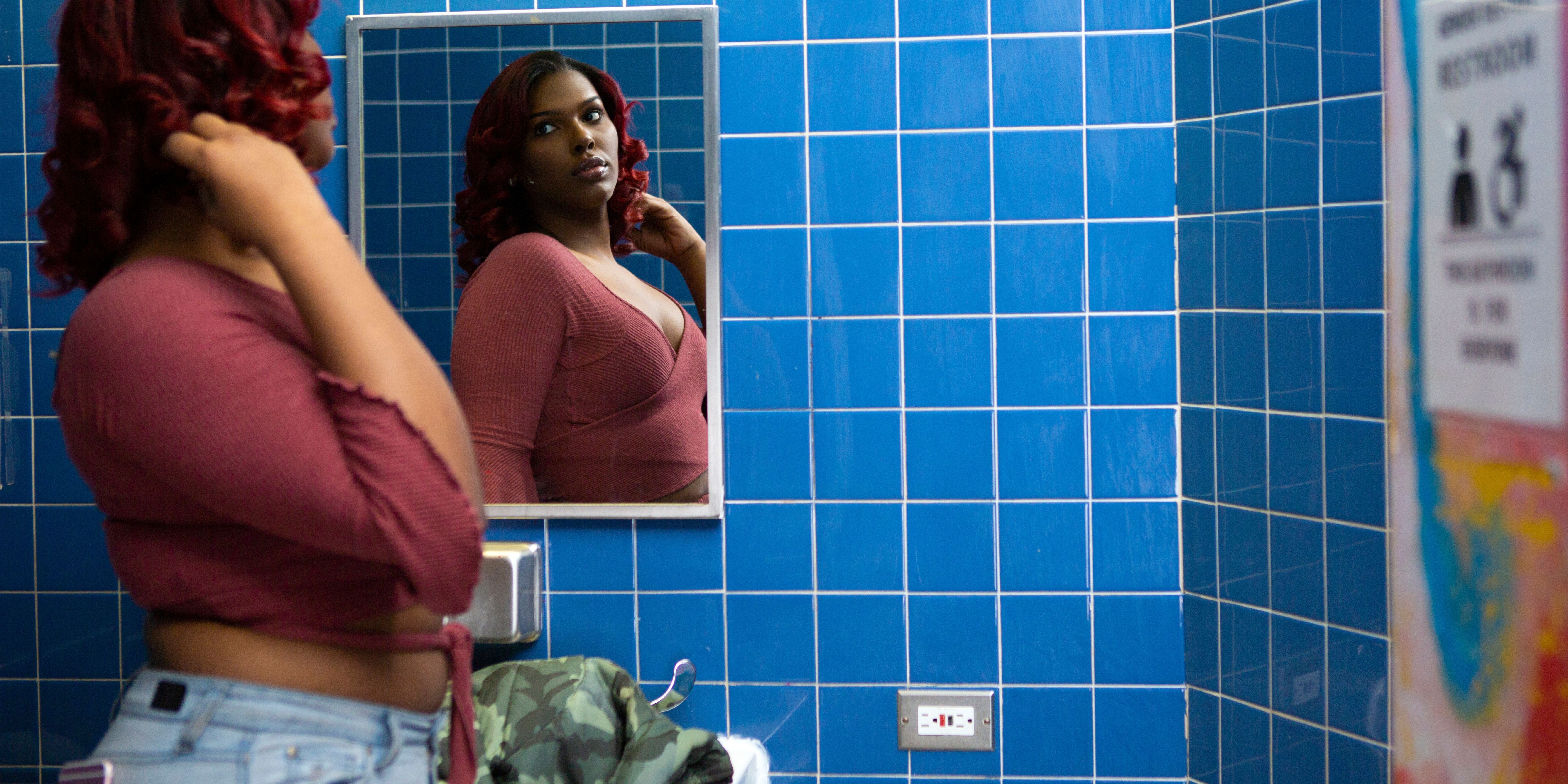 A dark-skinned transgender woman looking in the bathroom mirror at school.