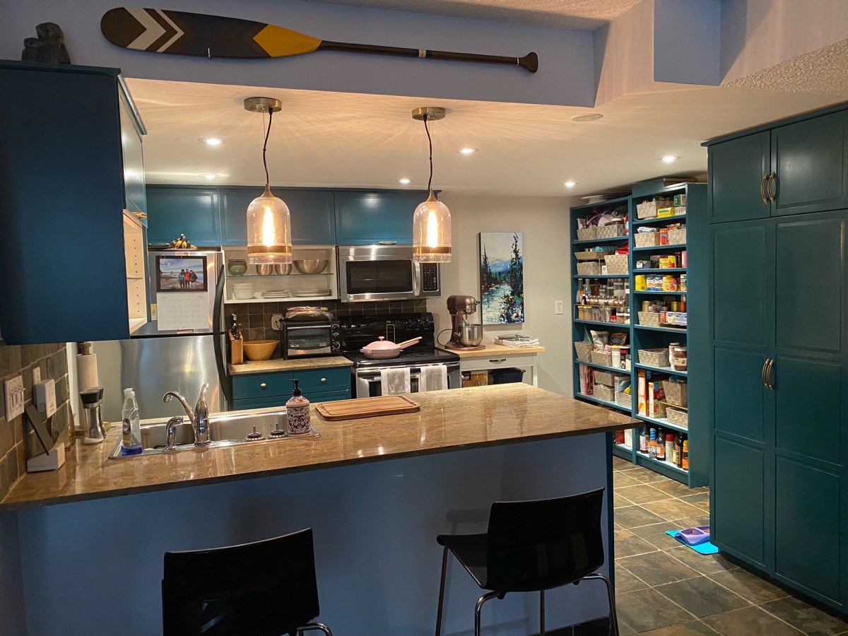 A blue and turquoise kitchen with warm brown counter tops, an oar mounted on one of the walls, and a large pantry shelf visible on the right side. Pink tools and pans sit on the counter as well as other cooking appliance