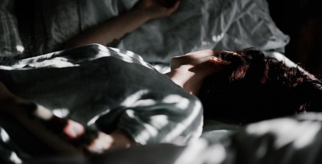 A dramatically lit photo of a person laying on their back in rumpled bedsheets, head turned to the side away from the camera