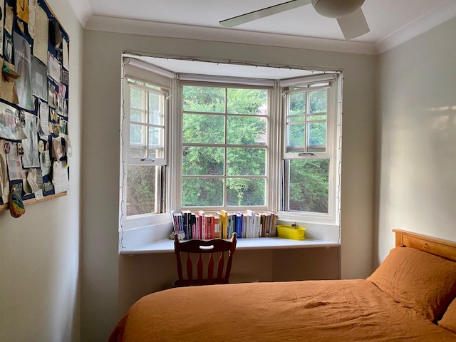 Large picture window with built in shelf, books lined up with a chair and office supplies, with bed in the foreground covered in orange quilt