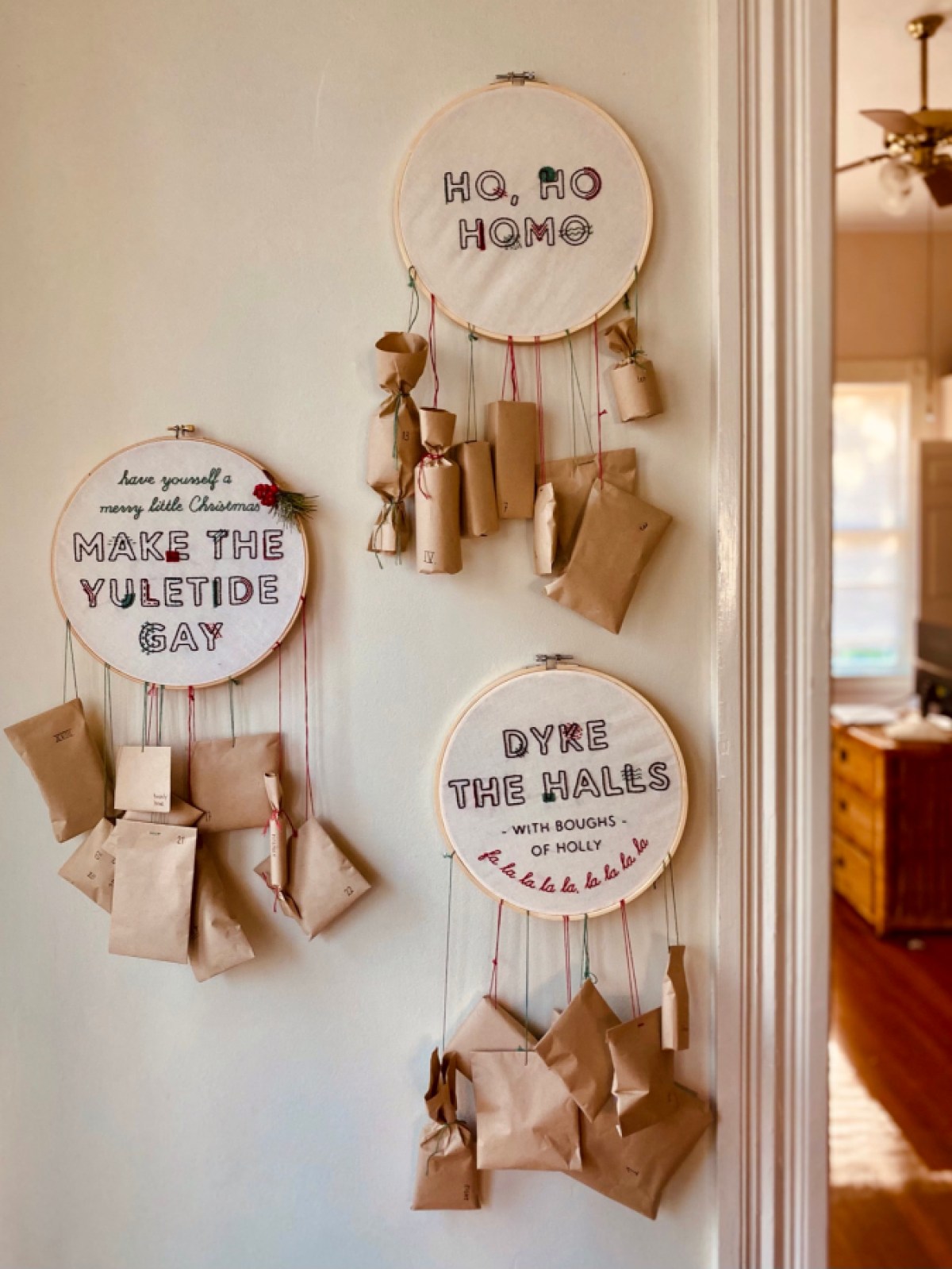 Three framed cross stitch pieces hang on a white wall, reading "Ho Ho Homo," "Have yourself a merry little Christmas, make the yuletide gay" and "Dyke the halls with boughs of holly." Each cross stitch has small brown paper wrapped parcels tied to the bottom