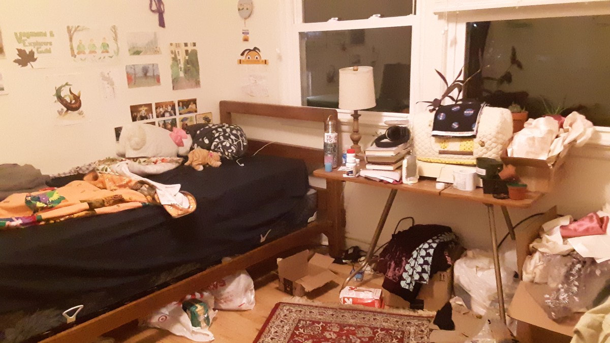 White walls with unframed prints hold a wooden bedframe covered in dark bedding, and a small table on the right side holds books, a vintage sewing machine, and other supplies