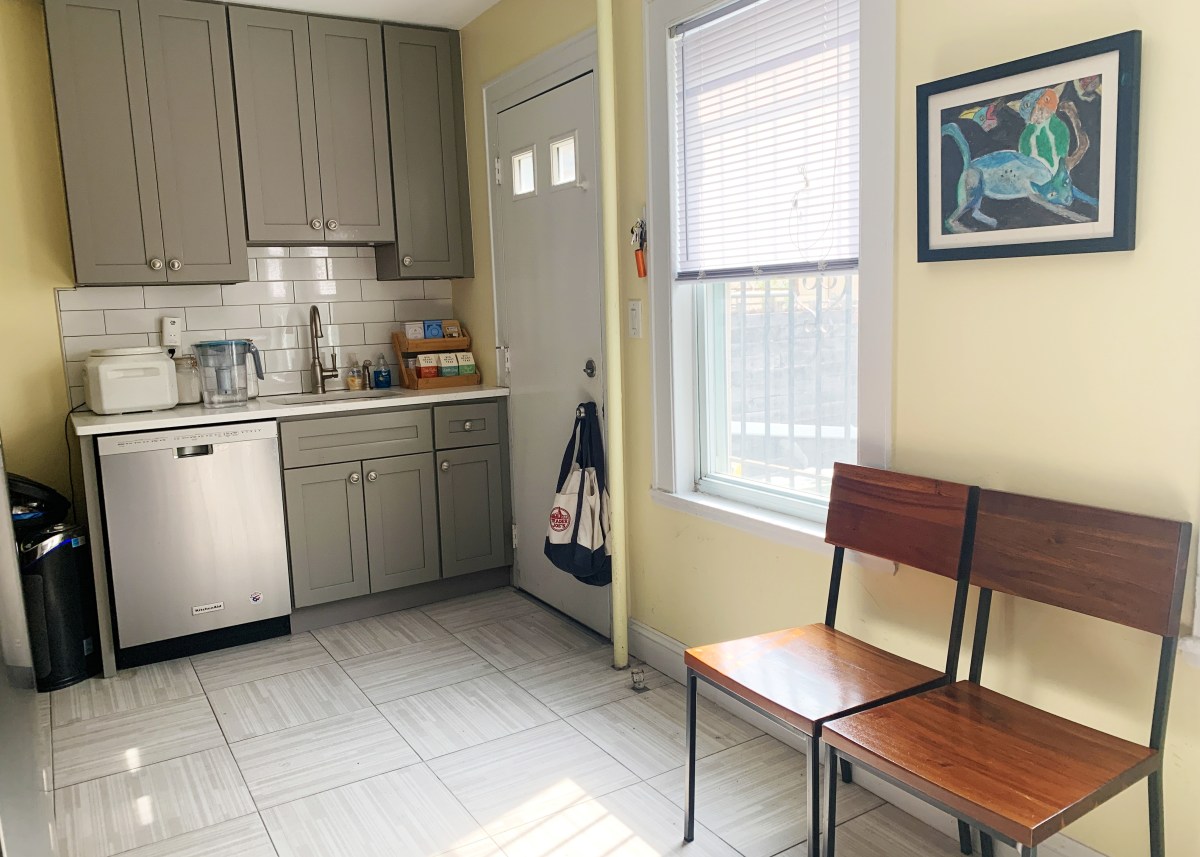 Kitchen with two dining chairs tucked against the right wall, cabinets and dishwasher visible as well as window with sun streaming through onto the floor.