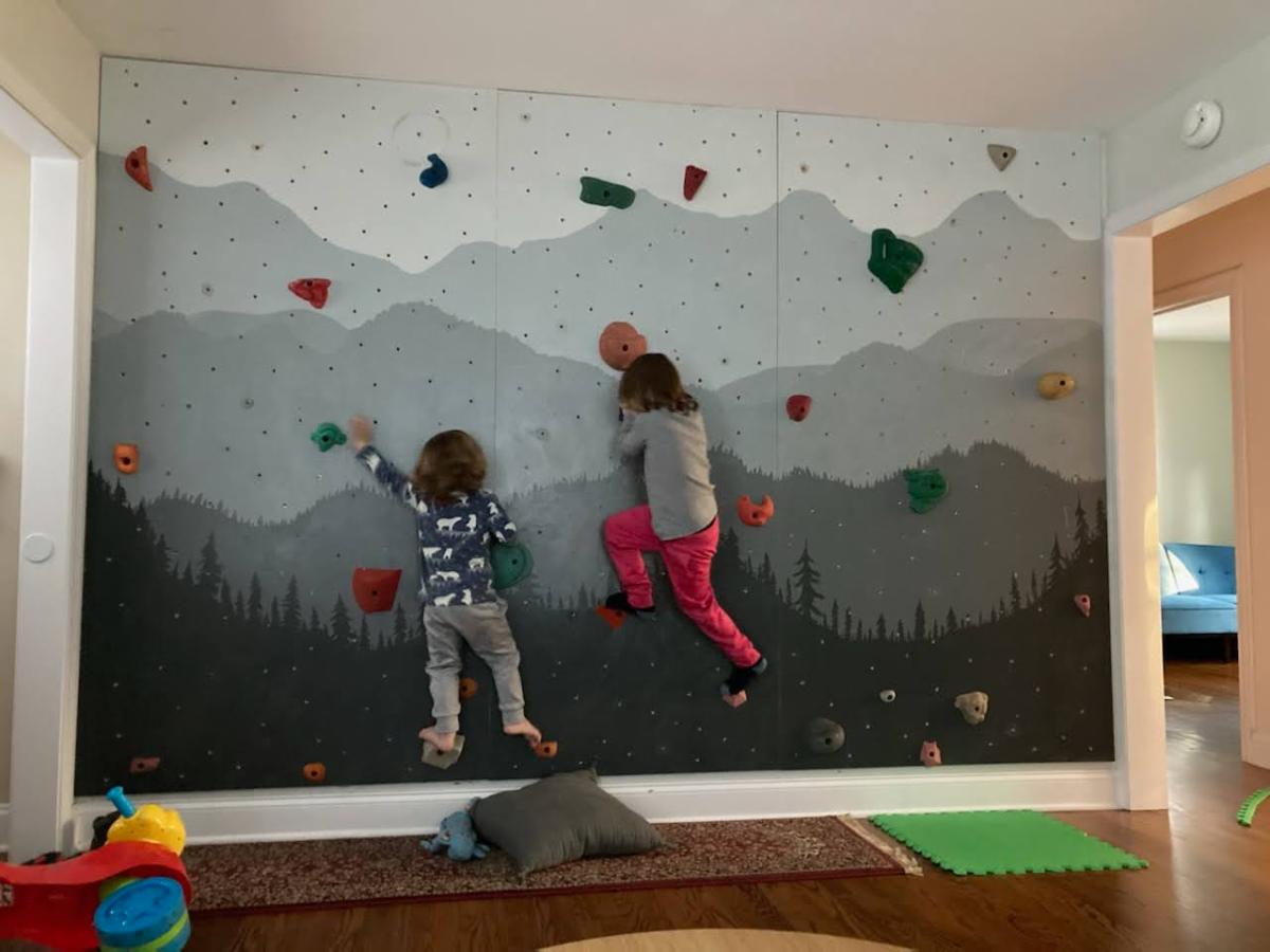 Indoor climbing wall with grey and blue mountains painted on it, two children are climbing up using the handholds