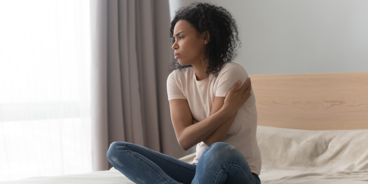 Black woman with curly medium length hair sits on a bed, wearing jeans and a white tee, hugging herself with her crossed arms, looking away from the camera as though in thought.