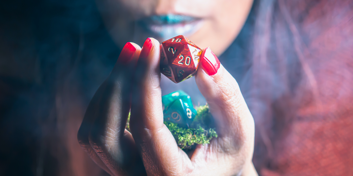 Image of a red 20 sided rpg die hold by a woman of color blowing onto it for good luck.