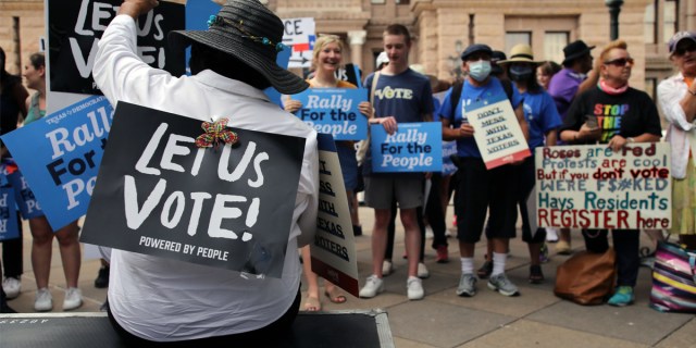 Photo from a Texas protest against Texas SB 7, a bill that would place new restrictions on access to the polls, where a person in the foreground raises their fist with a sign strapped to their torso that reads LET US VOTE