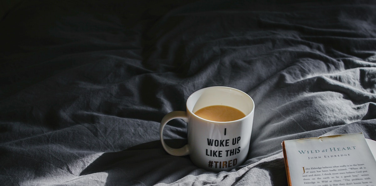 A dramatically lit photo of a cup of coffee in a mug that reads "I woke up like this #tired" nestled into rumpled bedsheets along with an open hardcover book.