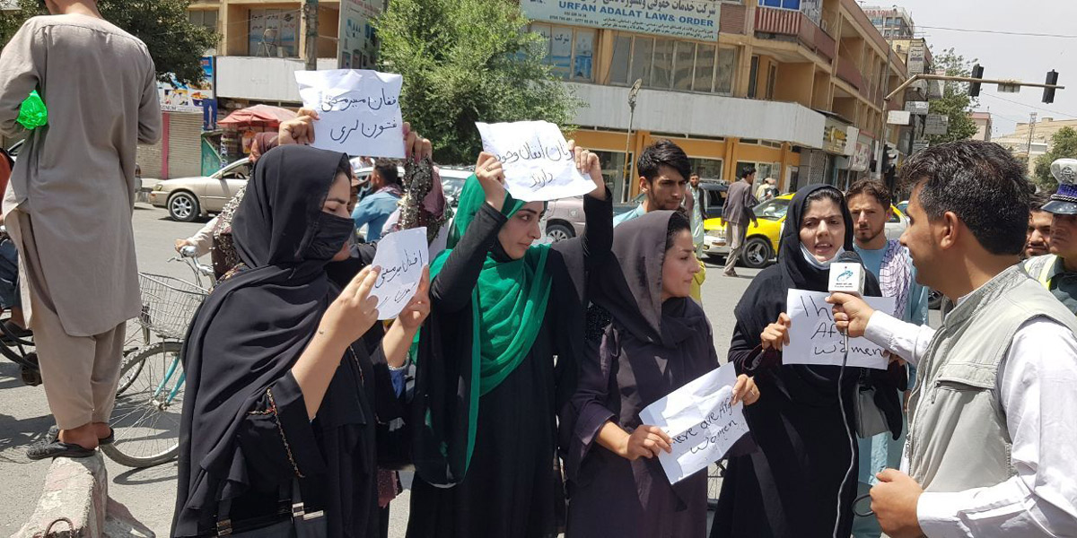 A group of Afghan women, one in a green hijab holding a sign, are interviewed by a reporter in Afghanistan.