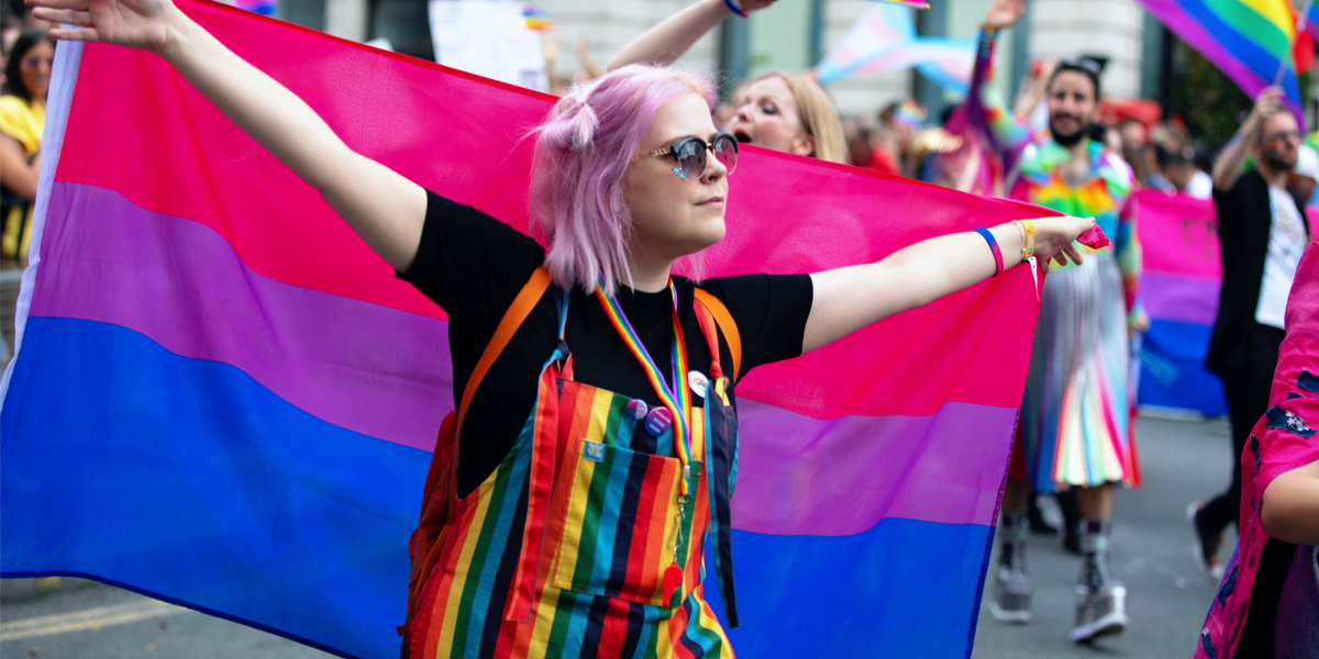 A person with shoulder length pink hair and rainbow overalls uses the bisexual pride flag as a cape during a pride march.