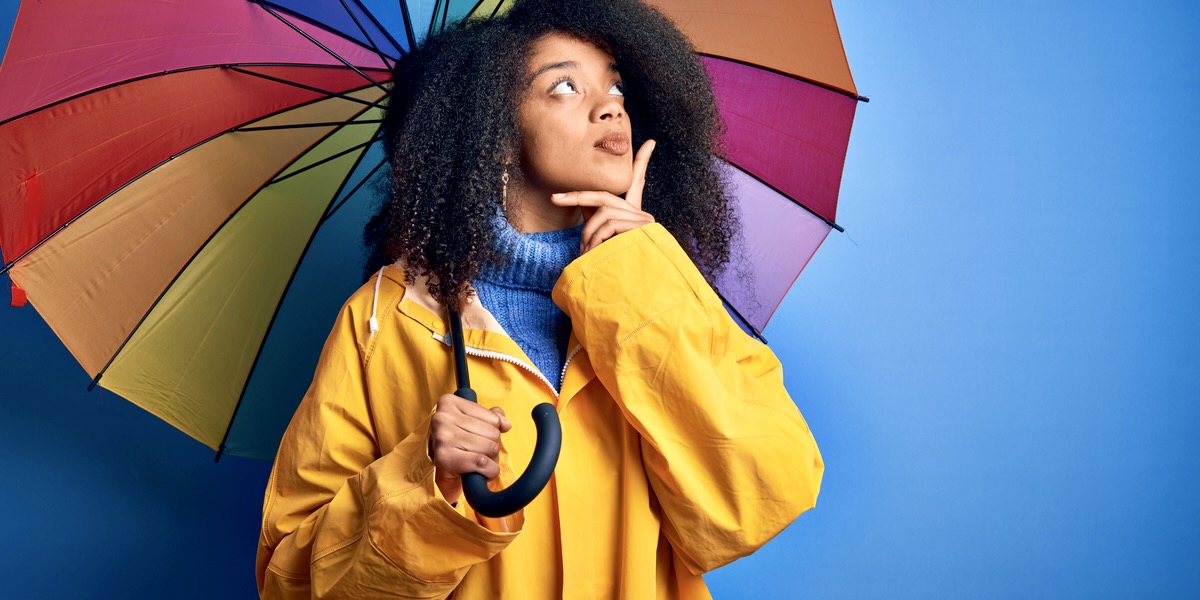 A black woman with curly hair has an inquisitive looks off to the right while pondering what does the word sapphic mean underneath a rainbow umbrella.