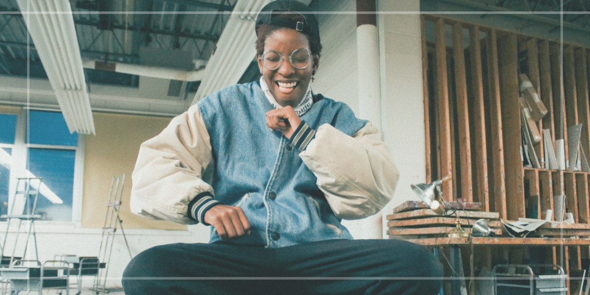 Stud Lesbian — Image shows a Black person with a joyful smile mid dance wearing a oversized vintage denim jacket, a backwards hat, round glasses and black pants. They are sitting down and again, joyfully mid dance.