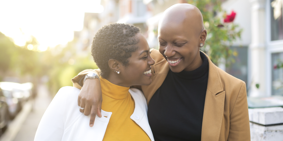 A Black person with a shaved head wearing a black shirt and brown blazer throws their arm around another Black person with short hair wearing a yellow shirt and white jacket.