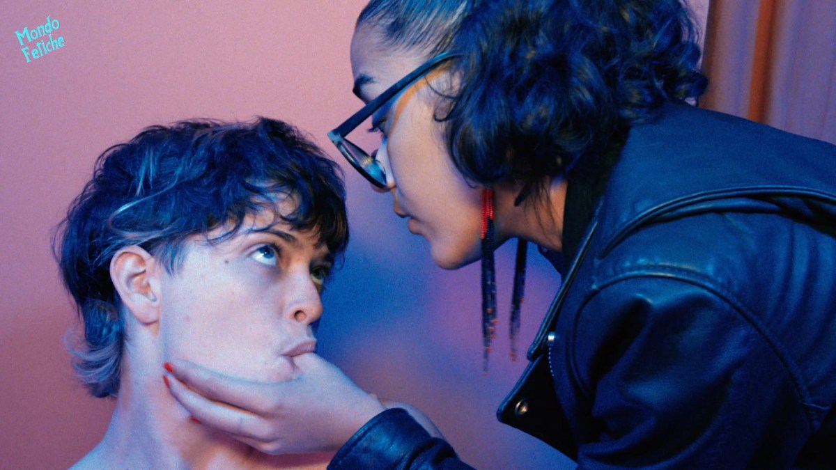 Image shows 2 people looking into each others eyes. One person wearing a leather jacket has their thumb in the other persons mouth.