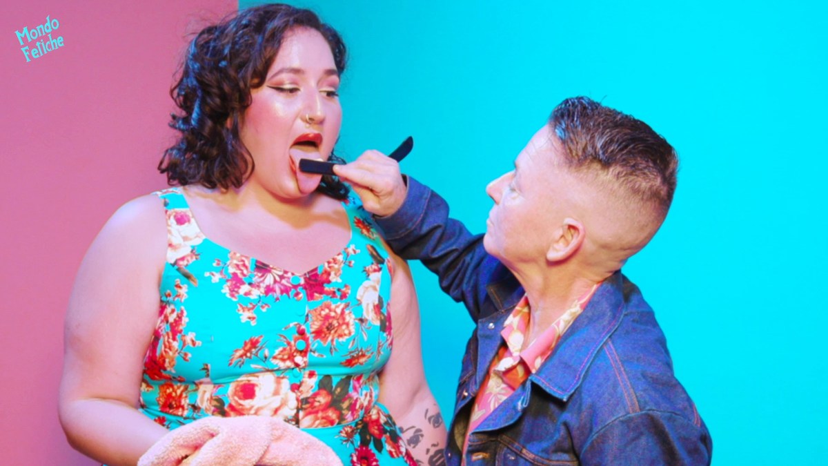 Image shows a pin-up styled person with her tongue out, while another person wearing a denim jacket puts their shaving razor on the others tongue making her lick it clean.