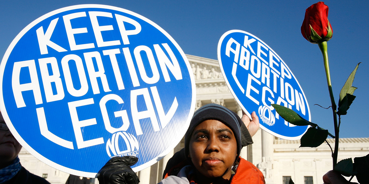 Today is a good day to donate to an abortion fund: A black person holds a large blue sign in the sky that says "Keep Abortion Legal."