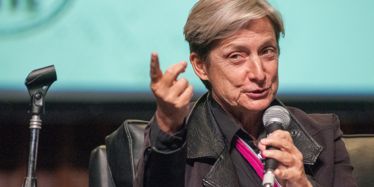 Judith Butler talks into a microphone against a blue background, they are wearing a pink tie.