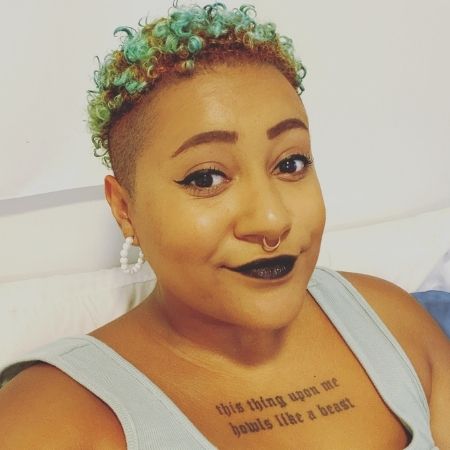 Jade, a person with short, curly blue hair that's shaved on the sides, wears black eyeliner, black lipstick and a septum piercing. They have a tattoo of text across their chest.