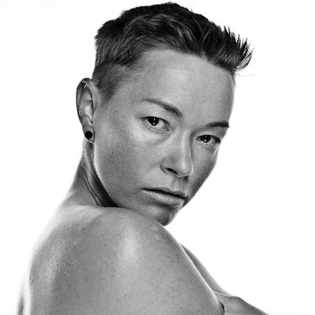 A black and white image shows Jiz Lee, a shirtless person with short hair, crossing their arms and turning over their shoulder.