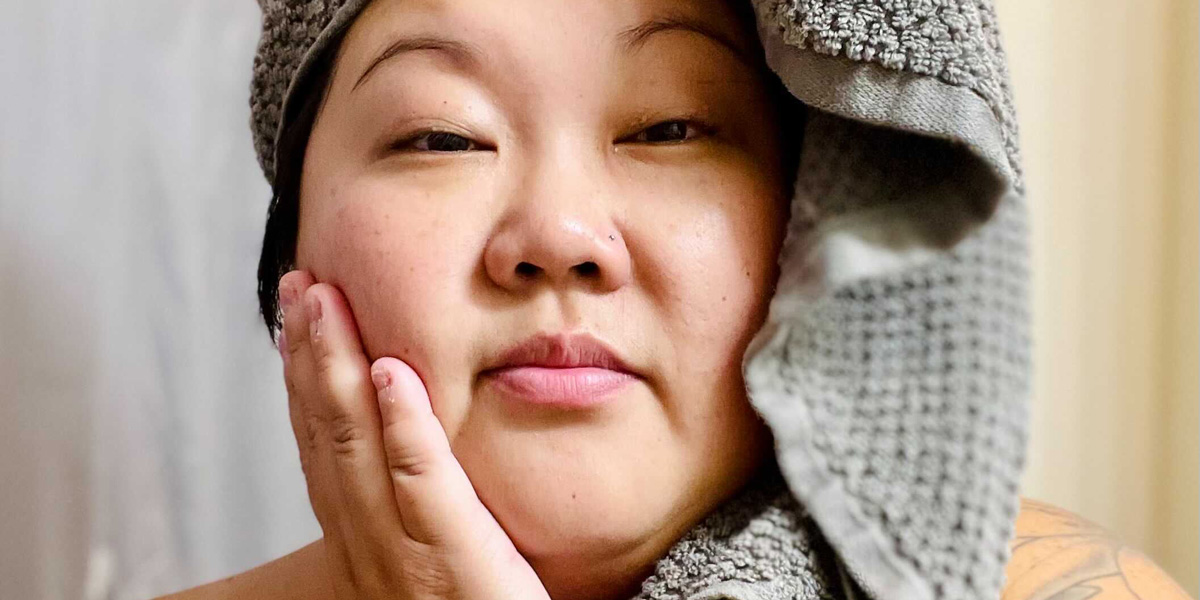 A femme Asian woman in her 30s is fresh faced from the shower and has a towel wrapped around her face.