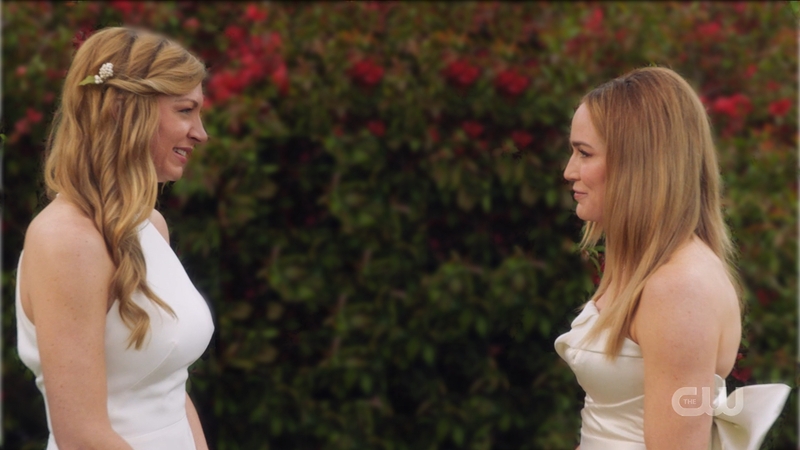 Ava and Sara stand in their wedding outfits smiling at each other.
