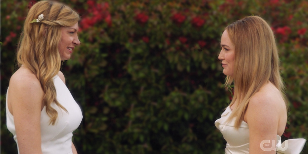 Ava and Sara stand in their wedding outfits smiling at each other.