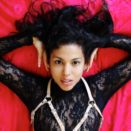 Lina Bembe is on against a red background wearing a black lace top and a white chest harness