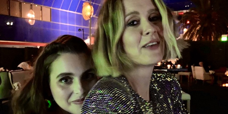 In the middle of a dark club, Beanie Feldstein hugs Sarah Paulson from behind while Sarah looks seductively into the camera.
