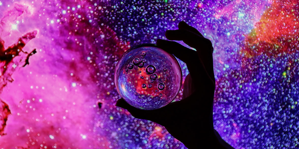 A hand that is backlit holds a magnifying glass up against a galaxy that is lit up in bisexual lighting.