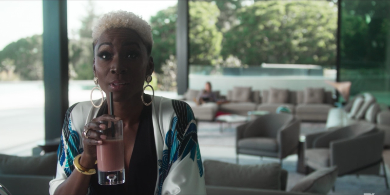 Angelica Ross with short blonde hair, a white silk robe, and gold earrings drinks from a smoothie while watching TV.