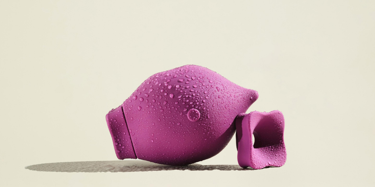 The Poet, a magenta suction sex toy shaped like a tear drop, is against an off-white background. It rests against its detachable suction mouth piece. The toy is slightly wet.