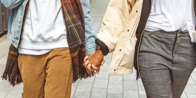 A person wearing a white sweatshirt, a plaid scarf, a denim jacket and tan corduroy pants holds hands with a person wearing a white shirt, a cream-colored jacket and plaid pants.