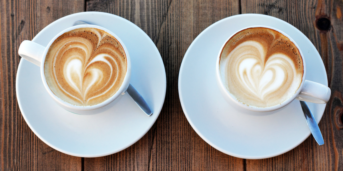 Two white coffee cups are on white plates with spoons. Each cup features latte art of a heart.