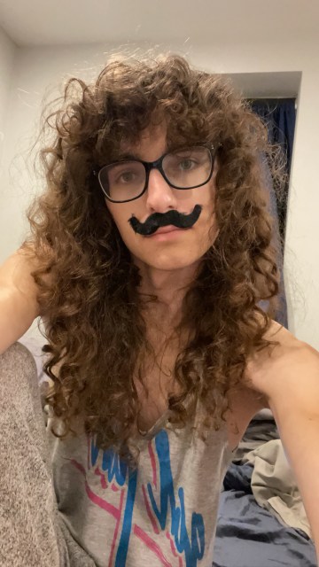 Drew, Autostraddle writer, takes a selfie while wearing the Autostraddle Gal Pal tee that she's cut the sleeves off. Drew has pasted a fake costume mustache onto her face. 