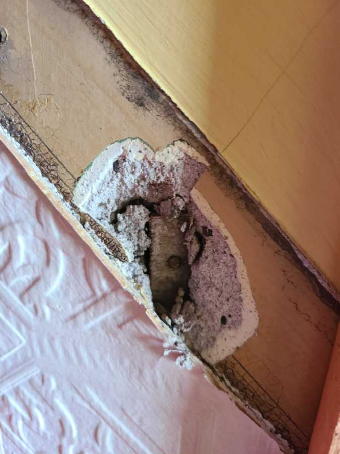 A photograph of a little hole in the wall of a stairway with a tiny bit of wooden lath with a nail in it peeking out from behind plaster.