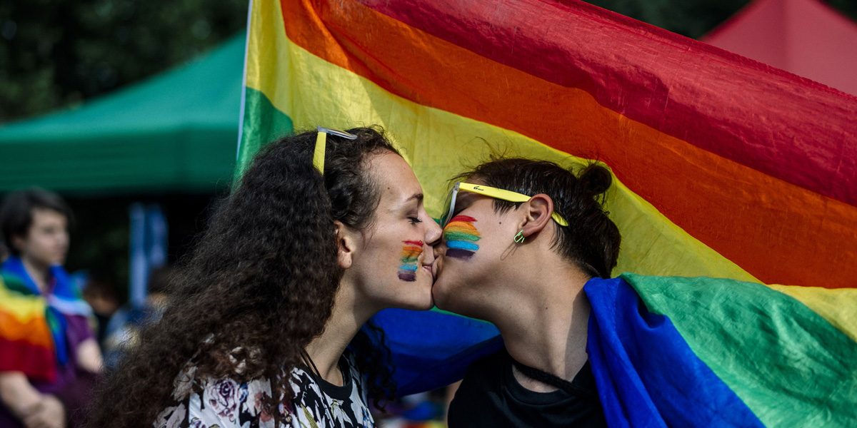Two femme queers kiss in front of the pride flag while also having rainbow hearts painted on their faces