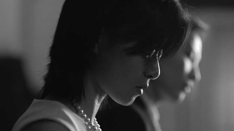 A black and white close up of a woman's face as she looks down at a piano. Another woman sits next to her out of focus.