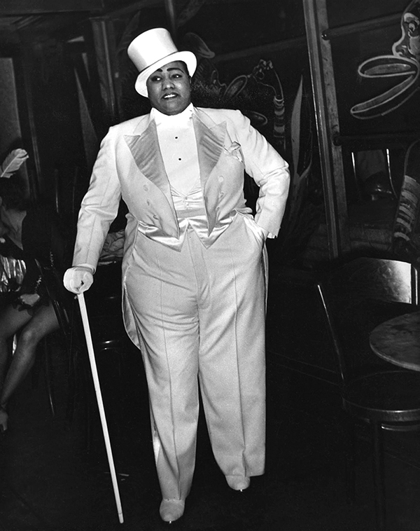 Gladys Bentley stands majestically in an all-white tuxedo and top-hat, leant on a white cane