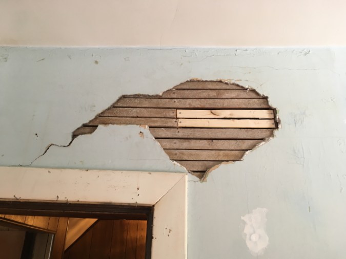 A look at a piece of wall above a doorway. A good chunk of plaster has fallen away revealing the lath. Two pieces of lath are a much brighter color than the other lath indicating their newness.