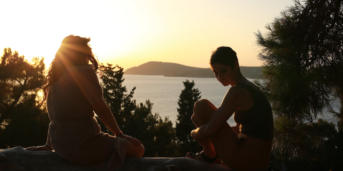 Selen Uçer as Reyhan and Ece Dizdar as Eren sit by the water as the sun sets