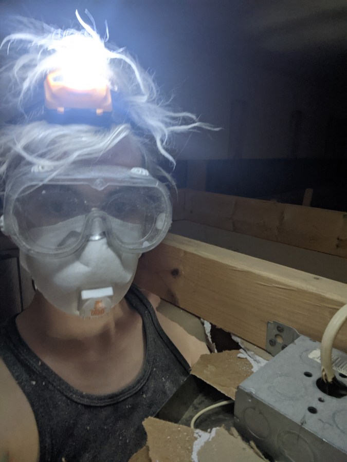 Nicole in a hole. Nicole's head and shoulders poke up through a hole in the second ceiling installed beneathe the original ceiling. They are wearing a lit headlamp, glasses, goggles and an N95 mask and tank top. They are a genderqueer white human with bleached hair.