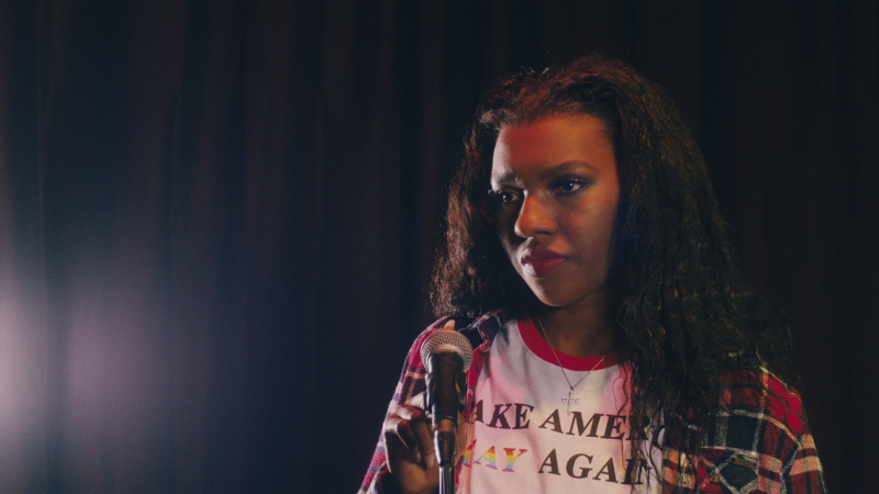 A Black teenage girl stands at a microphone wearing a Make America Gay Again shirt and a flannel.