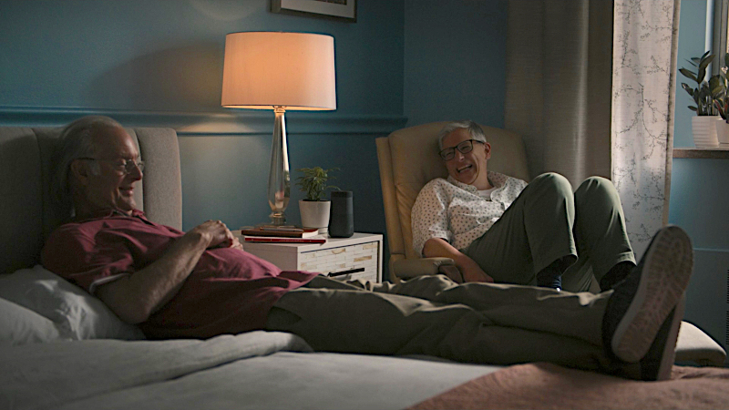 Abby sits in a chair next to her dad who is lying on a bed. They are both laughing.