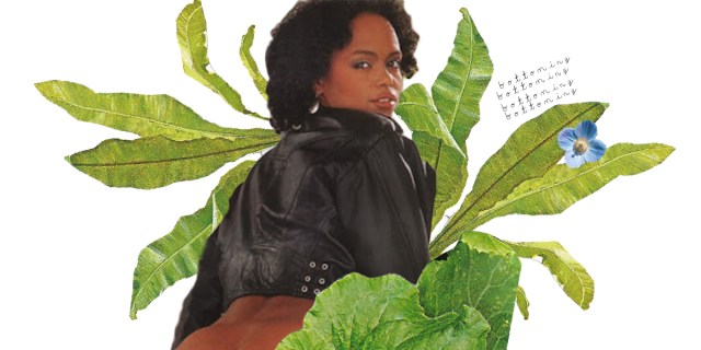 A Black woman in a black leather jacket is surrounded by green leaves and a small blue flower